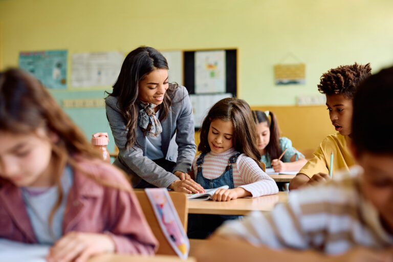 Happy elementary school teacher assisting her student in the classroom.