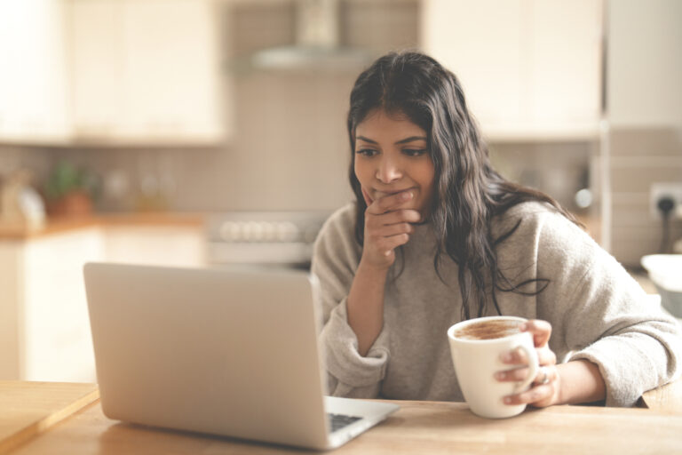 A young woman, deep in thought, enjoying her coffee as she navigates her laptop in a warm and inviting kitchen. The sunlit space creates a tranquil atmosphere.